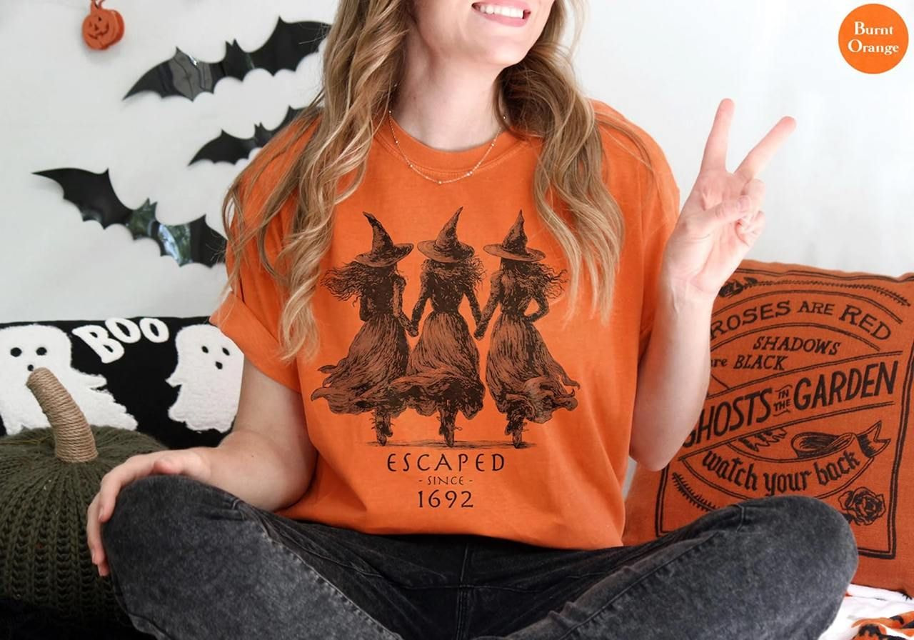 A woman wearing an orange Halloween-themed shirt with three witch silhouettes and "Escaped Since 1692" text.
